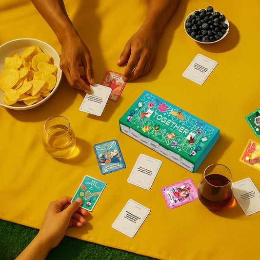 Three people play the Together Couples Intimacy Card Game on a yellow blanket, with bowls of chips, blueberries, and drinks beside them. They hold and draw cards designed to spark deeper conversations and strengthen their relationship.