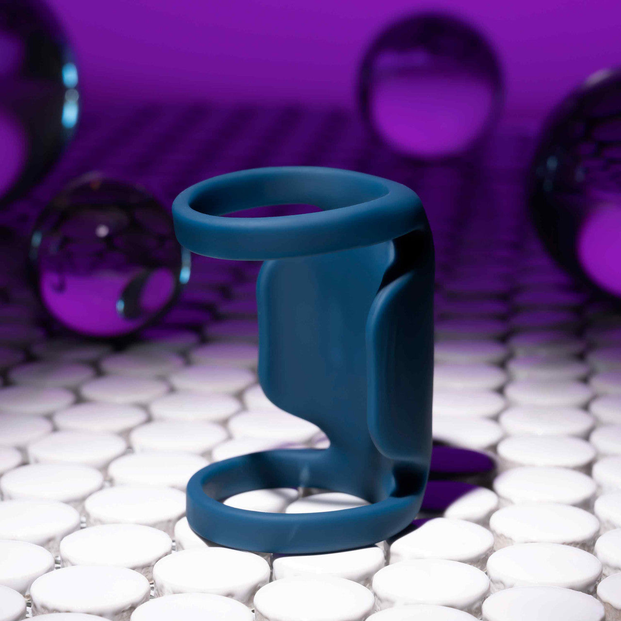 A modern blue 3D object, identified as the Jet Pack Vibrating Penis Enhancer Sheath by Evolved Novelties, and crafted from body-safe silicone, rests on a white tiled surface. The purple background features reflective spheres scattered around, forming a futuristic and abstract scene.