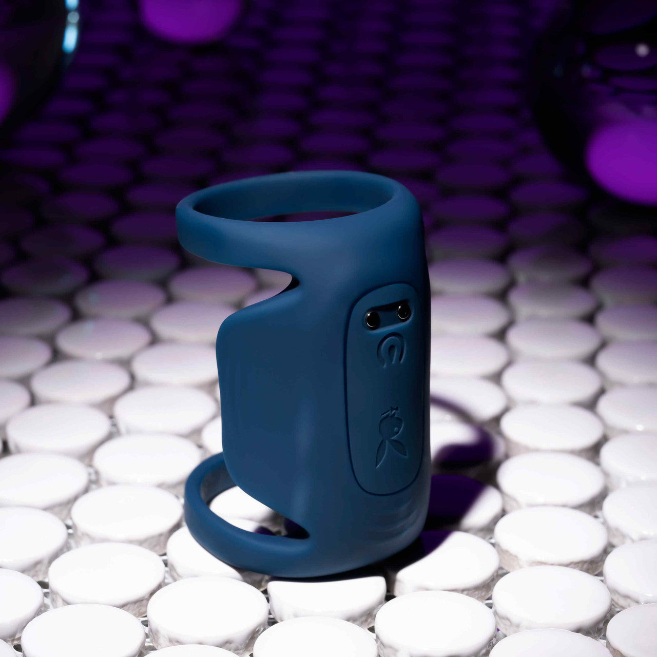 A blue ring-shaped device, the Jet Pack Vibrating Penis Enhancer Sheath from Evolved Novelties, crafted from body-safe silicone, sits elegantly on a tiled surface. The background, illuminated with purple lighting, casts a modern and futuristic ambiance, making it feel as innovative as the USB rechargeable technology it discreetly incorporates.