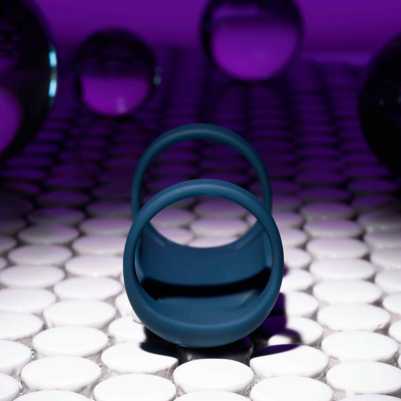 A Jet Pack Vibrating Penis Enhancer Sheath by Evolved Novelties, crafted from body-safe silicone, sits on a reflective surface adorned with a pattern of white circular tiles. The background is bathed in a purple glow, with several glass spheres softly blurred in the distance to create an air of mystique.