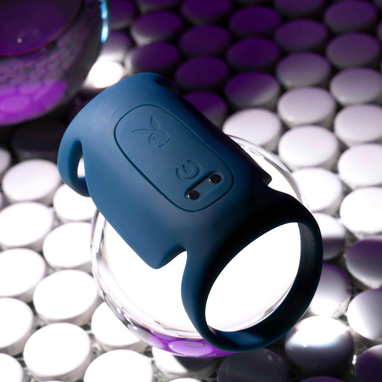 A Jet Pack Vibrating Penis Enhancer Sheath by Evolved Novelties, designed in blue body-safe silicone with a rabbit motif and button feature, is displayed elegantly on a glass orb amidst white circular tiles, all bathed in purple lighting.