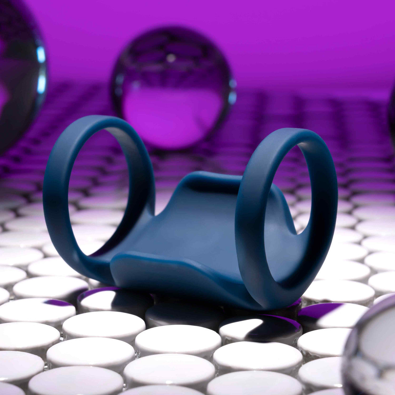 A Jet Pack Vibrating Penis Enhancer Sheath by Evolved Novelties, featuring two circular handles and crafted from body-safe silicone, rests on a reflective white tiled surface. The background is purple with reflective spherical objects scattered around.