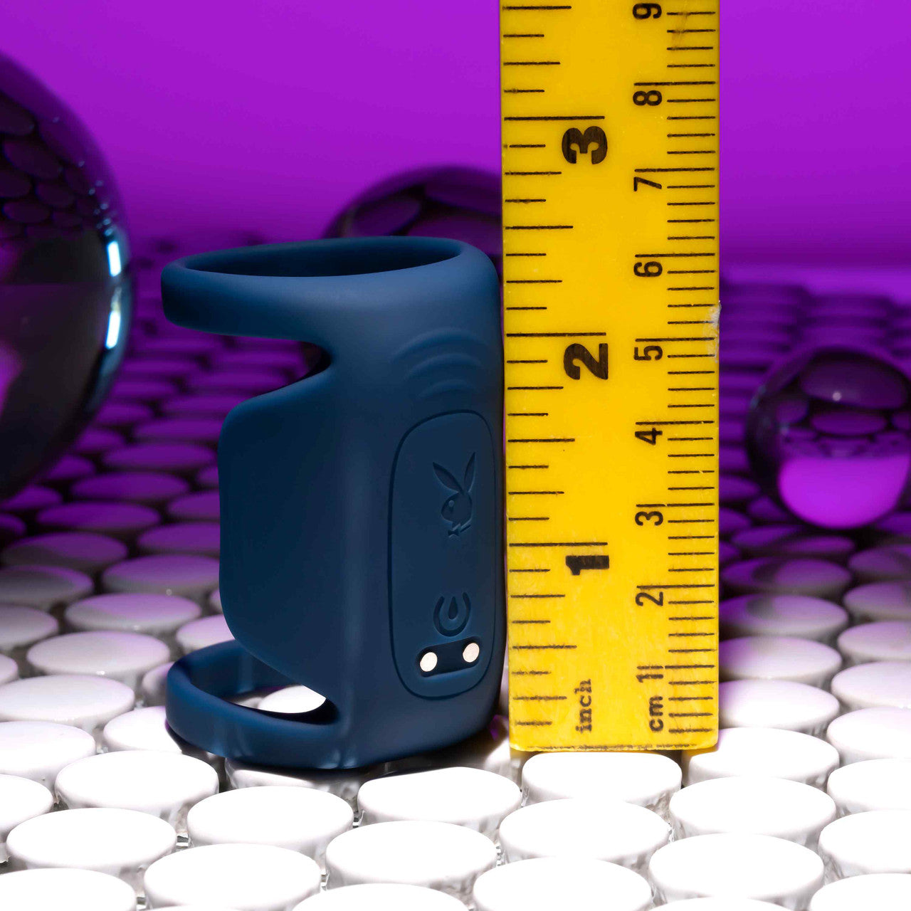 The Jet Pack Vibrating Penis Enhancer Sheath by Evolved Novelties, crafted from body-safe silicone and integrated with electronic components, is positioned beside a yellow measuring ruler that marks its height at approximately 2 inches. Against a backdrop of reflective white tiles enhanced by purple lighting, it boasts a stylish appearance and USB rechargeable functionality.