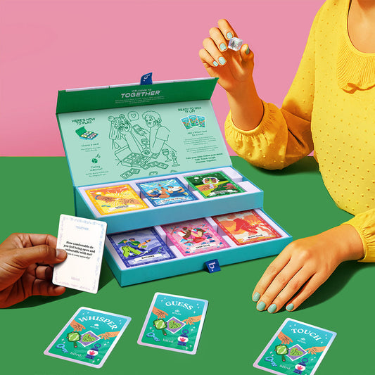 A person in a yellow top holds a die next to the open Together Couples Intimacy Card Game box with illustrated cards and dice. Another hand has a white card. Cards labeled "Whisper," "Guess," and "Touch" inspire communication for couples.