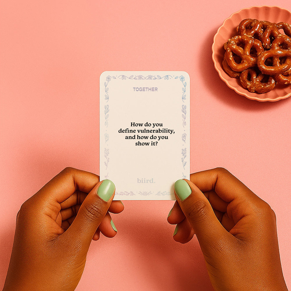 Two hands with mint green nail polish hold a Together Couples Intimacy Card Game card that says, "How do you define vulnerability, and how do you show it?" A pink bowl of pretzels sits on a pink surface in the background.