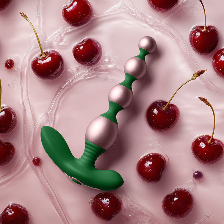 The Bougie Vibrating Aluminum Anal Beads in green and pink rest on a glossy pink surface, surrounded by fresh cherries and cherry juice.