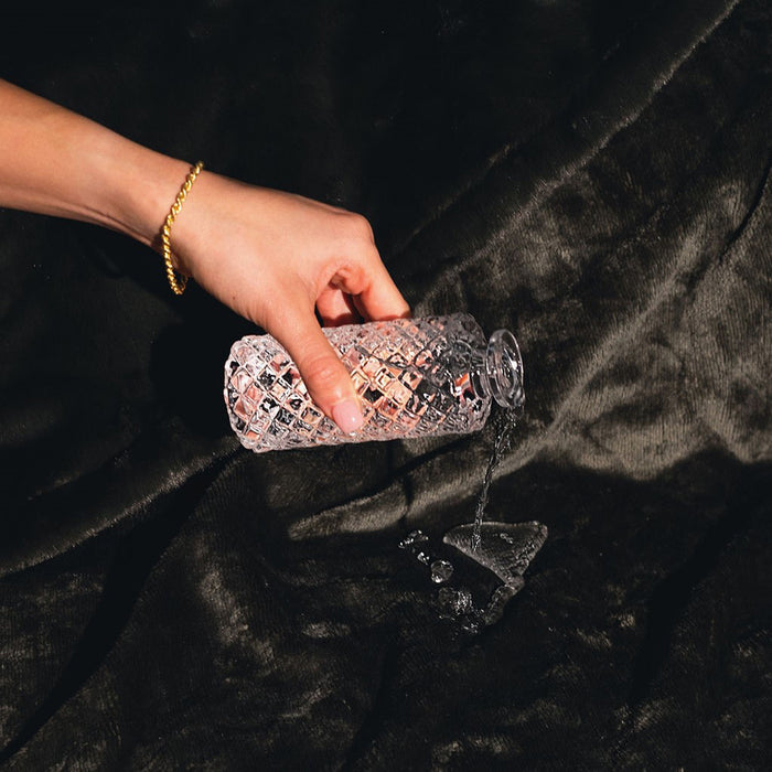 A hand pours water from a patterned glass bottle onto a Splash Blanket Furlush Luxurious Faux Fur Waterproof Blanket - Small, where splashes form a puddle on the plush, dark, textured surface.