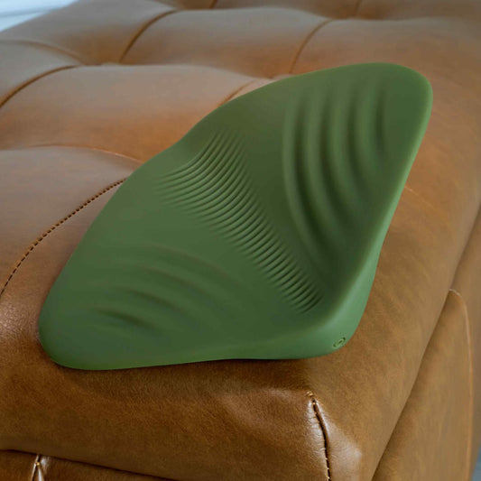 A green Gender X Pleasure Pad Rechargeable Grinding Pad & Stroker with textured ridges sits on a tan leather tufted ottoman.