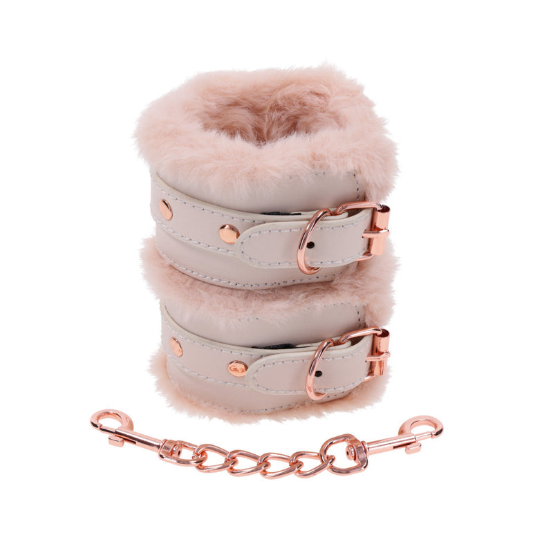 A pair of pink, fluffy Peaches 'n Cream Fur Handcuffs from Sportsheets, featuring rose gold buckles, are stacked together. Below is a short chain with clips on both ends that match the buckles. These bondage accessories are elegantly showcased on a plain white background.