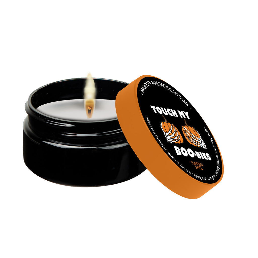 A black container candle with a lit wick is shown. The lid, resting to the side, reads "TOUCH MY BOO-BIES" in white text on an orange background. It also says "WARM TOUCH MASSAGE CANDLES" and displays images of pumpkins, hinting at the Pumpkin Spice Erotic Massage Candle by Kama Sutra for an erotic massage experience.