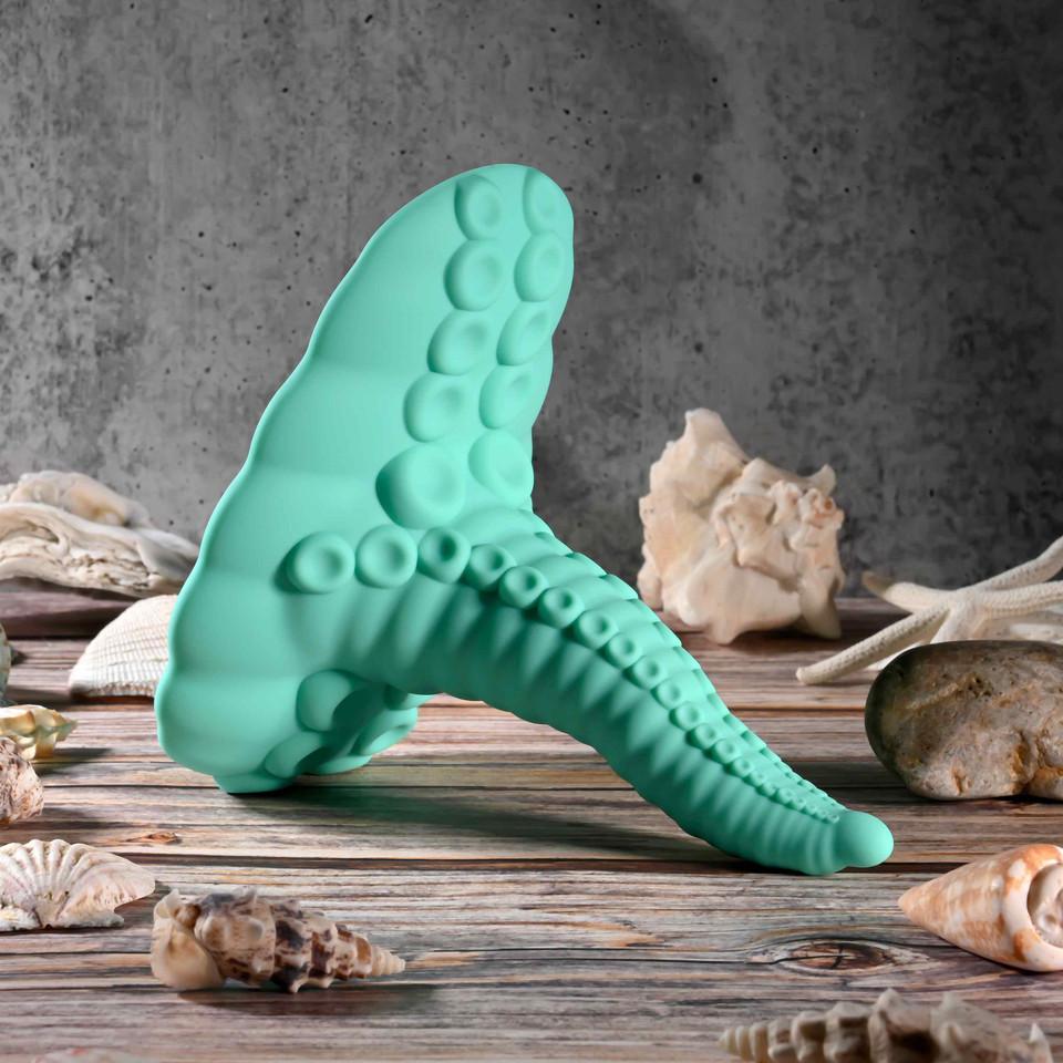 The Octopus Tentacle Insertable Remote Controlled Grinder, a green silicone vibrator shaped like an octopus tentacle, stands upright on a wooden surface among seashells and rocks with a gray textured wall in the background.