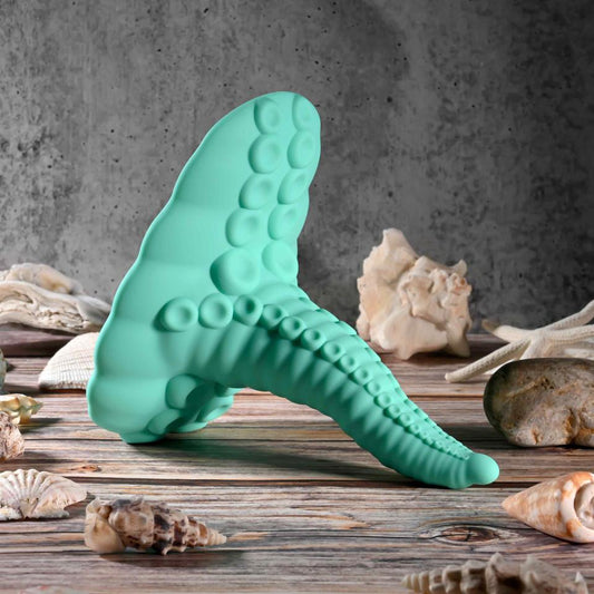 The Octopus Tentacle Insertable Remote Controlled Grinder, a green silicone vibrator shaped like an octopus tentacle, stands upright on a wooden surface among seashells and rocks with a gray textured wall in the background.