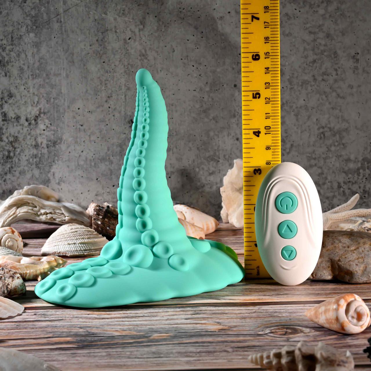 The Octopus Tentacle Insertable Remote Controlled Grinder—a teal, silicone, tentacle-shaped waterproof sex toy with suction cup details—is shown beside a measuring tape, white three-button remote, and seashells on a wooden surface.