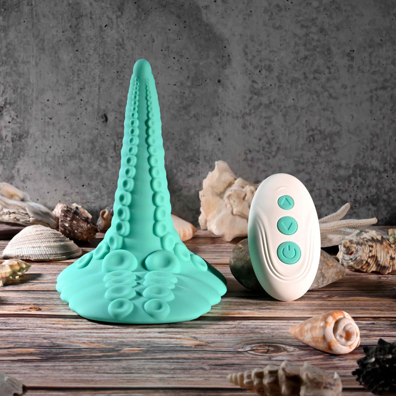 The Octopus Tentacle Insertable Remote Controlled Grinder, a teal waterproof toy, stands upright with its white remote among seashells on a wooden surface, set against a gray stone background.
