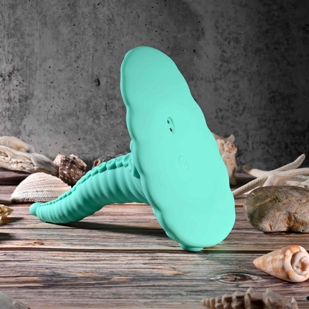 The Octopus Tentacle Insertable Remote Controlled Grinder, a turquoise silicone vibrator with a ribbed handle and cloud-shaped top, rests on wood amid seashells and coral against a textured gray wall.