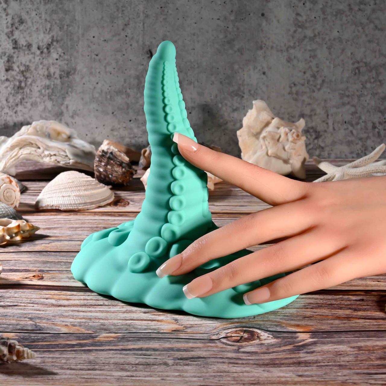 A realistic silicone hand touches the Octopus Tentacle Insertable Remote Controlled Grinder, a teal waterproof sex toy, on a wooden surface with assorted seashells and a gray stone background.